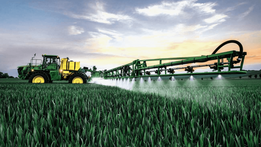 Large John Deere-style tractor with extended sprayer booms applying MaxxGro's bulk agrochemicals in India across a vast crop field, emphasizing high-volume supply chain capability.
