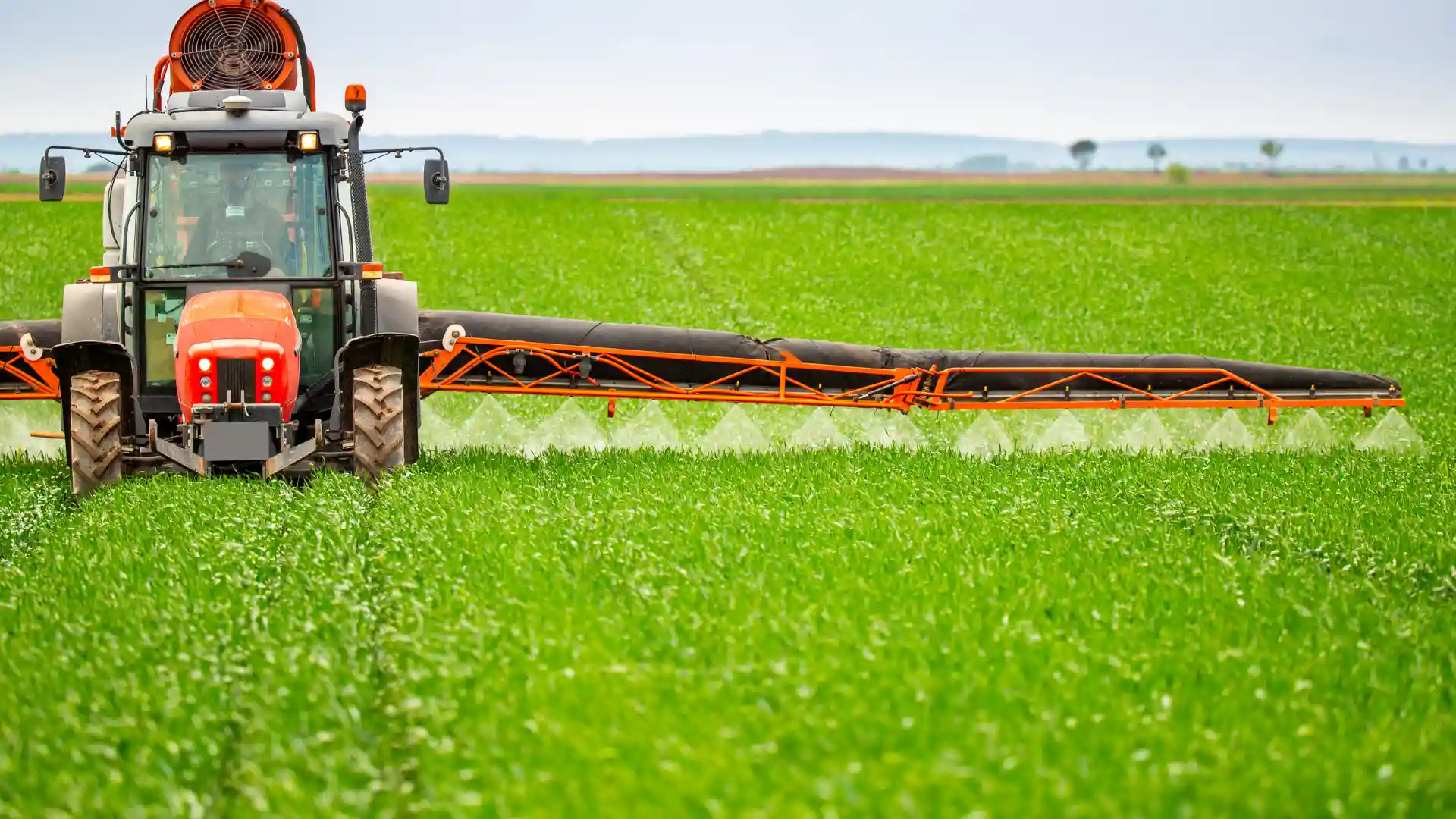 Modern agricultural tractor with a wide boom sprayer precisely applying agrochemical crop protection across a vibrant green field.