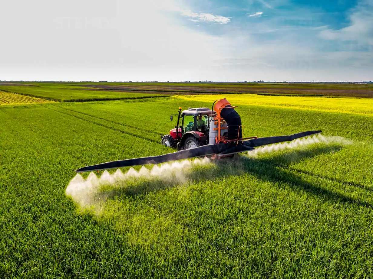 Aerial view of a self-propelled boom sprayer tractor applying agrochemical solutions across a massive, vibrant green field, emphasizing wide coverage and MaxxGro product efficiency.